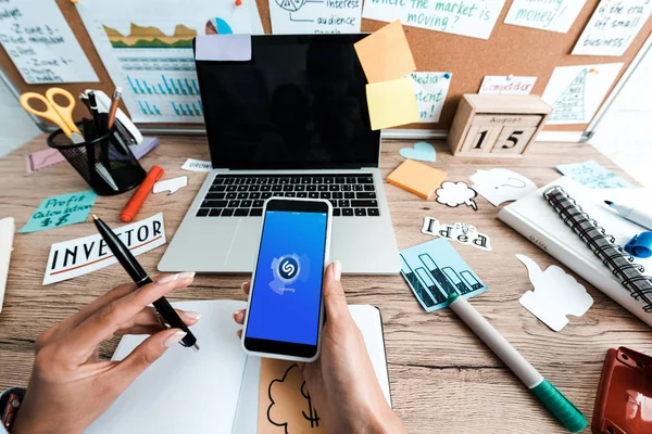 KYIV, UKRAINE - JULY 23, 2019: cropped view of woman holding smartphone with shazam app near sticky notes with lettering and laptop with blank screen — Stock Photo