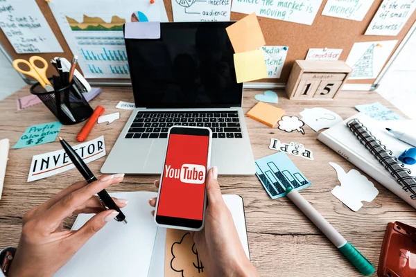 KYIV, UCRAINA - LUGLIO 23, 2019: vista ritagliata di donna che tiene smartphone con app youtube vicino a note adesive con lettering e laptop con schermo vuoto — Foto stock