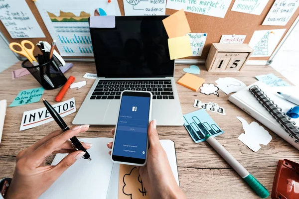 KYIV, UKRAINE - JULY 23, 2019: cropped view of woman holding smartphone with facebook app near sticky notes with lettering and laptop with blank screen — Stock Photo