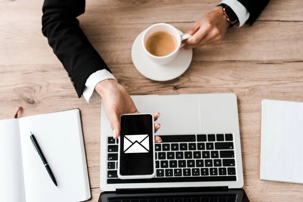 Vista dall'alto della donna d'affari che tiene smartphone con busta sullo schermo e tazza con caffè vicino al computer portatile — Foto stock