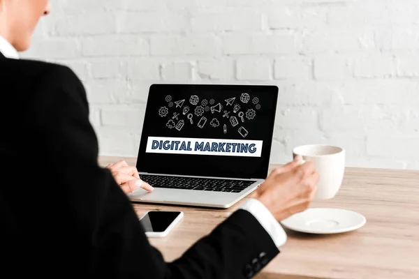 Selective focus of woman holding cup while sitting near laptop with digital marketing lettering and smartphone with blank screen — Stock Photo