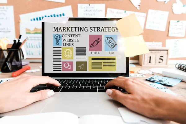 Cropped view of woman using laptop with marketing strategy lettering on screen near sticky notes — Stock Photo