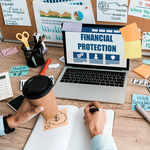 Vue recadrée de femme d'affaires avec stylo et tasse en papier près d'un ordinateur portable avec protection financière à l'écran — Photo de stock