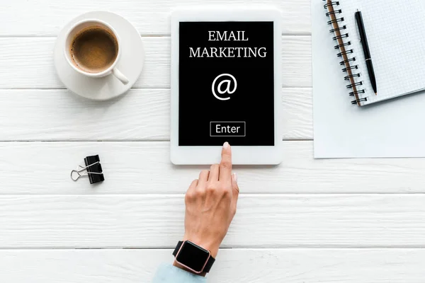 Ansicht von oben: Frau zeigt mit dem Finger auf digitales Tablet mit E-Mail-Marketing-Schriftzug auf dem Bildschirm neben Tasse Kaffee und Büroklammer — Stockfoto
