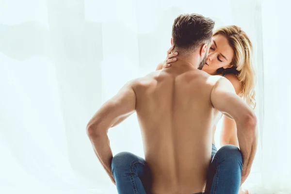 Young shirtless man kissing beautiful girlfriend by window at home — Stock Photo