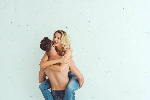 Vista trasera del hombre sin camisa sosteniendo las manos novia alegre mientras está de pie cerca de la pared blanca - foto de stock