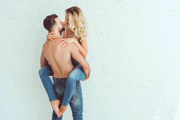 Vista posteriore di giovane uomo senza maglietta che tiene su mani bella fidanzata mentre in piedi vicino al muro bianco — Foto stock