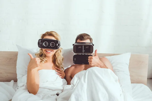 Young couple lying in bed in virtual reality headsets and showing thumbs up — Stock Photo