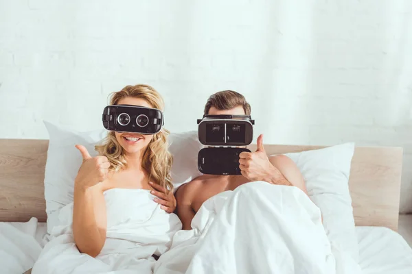 Pareja joven acostada en la cama en auriculares de realidad virtual y mostrando los pulgares hacia arriba - foto de stock