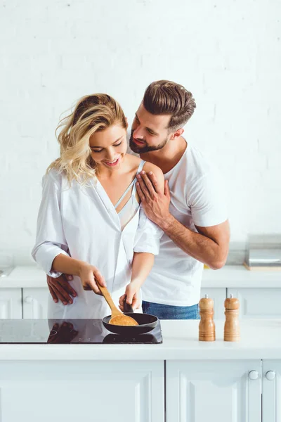Bonito homem abraçando bonita namorada preparando panqueca no frigideira — Fotografia de Stock
