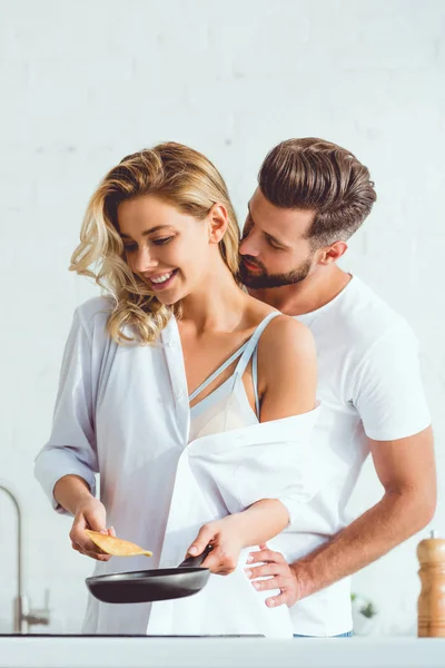 Guapo hombre abrazando feliz novia preparando panqueque en sartén - foto de stock