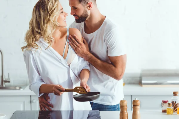 Bonito jovem abraçando alegre namorada preparando café da manhã na frigideira — Fotografia de Stock