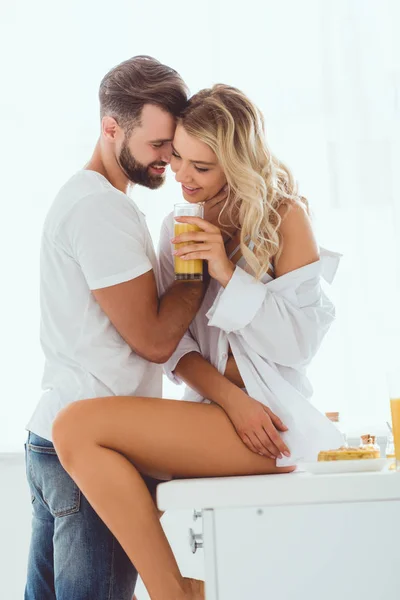 Feliz hombre abrazando bonita novia sentado en la mesa de la cocina y sosteniendo vaso de jugo de naranja - foto de stock