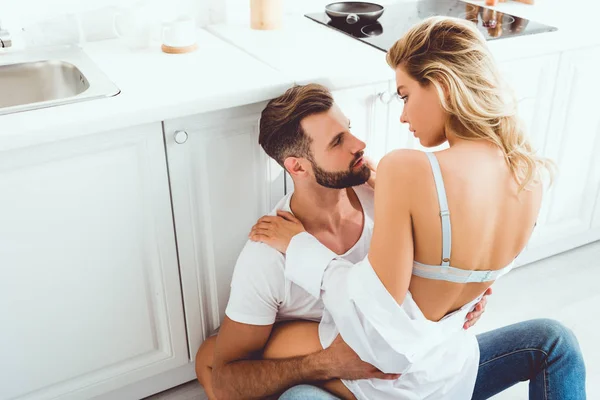 Pareja joven abrazándose y mirándose mientras están sentados en el piso de la cocina - foto de stock