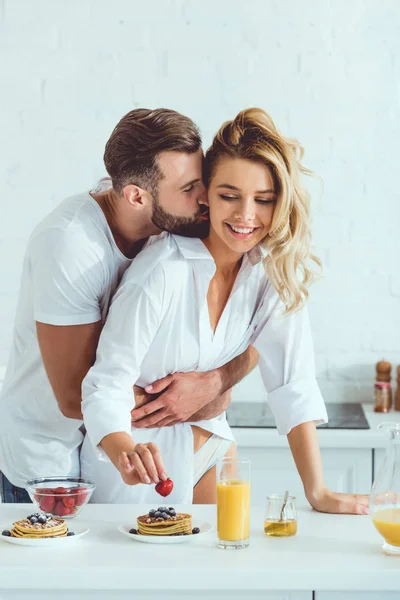 Guapo hombre abrazando y besando hermosa novia mientras que de pie cerca de la mesa con el desayuno servido - foto de stock