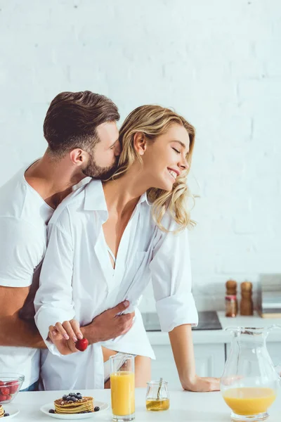 Joven abrazando feliz novia mientras está de pie cerca de la mesa de la cocina con el desayuno servido - foto de stock