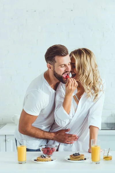 Happy young woman feeding boyfriend with strawberry while standing at kitchen table with served breakfast — Stock Photo
