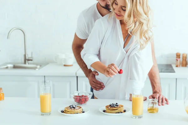 Vista recortada de un joven abrazando a una bonita novia mientras está de pie cerca de la mesa de la cocina con el desayuno servido - foto de stock