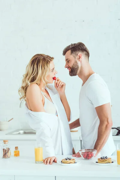 Heureux jeune homme debout près belle petite amie manger fraise dans la cuisine — Photo de stock