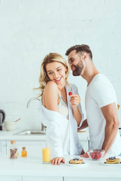 Sonriente hombre abrazando feliz novia sosteniendo fresa en cocina - foto de stock