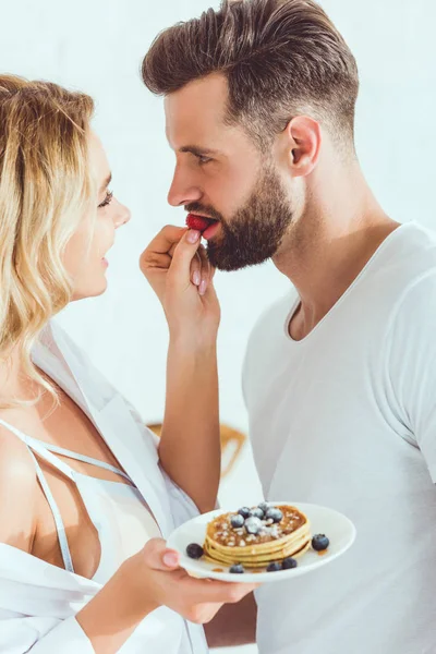 Giovane donna sensuale che alimenta il fidanzato con fragola mentre tiene il piatto con frittelle — Foto stock