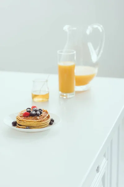 Plato de panqueques con bayas cerca de vidrio y jarra de jugo de naranja y tarro con miel sobre mesa blanca - foto de stock