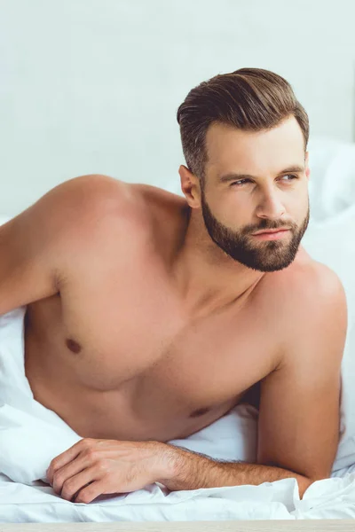 Pensativo, hombre sexy mirando hacia otro lado mientras está acostado en la cama en casa - foto de stock