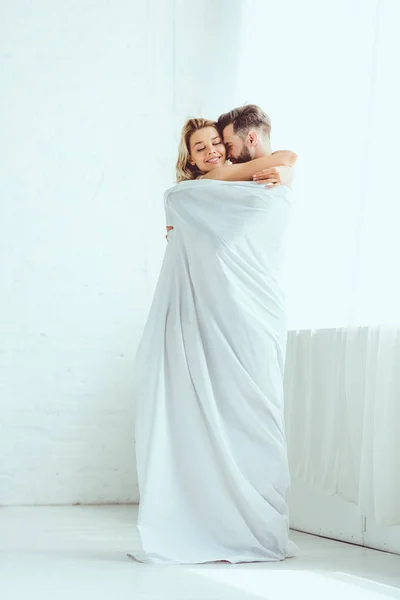 Счастливая молодая пара обнимается стоя завернутая в белую простынь — стоковое фото