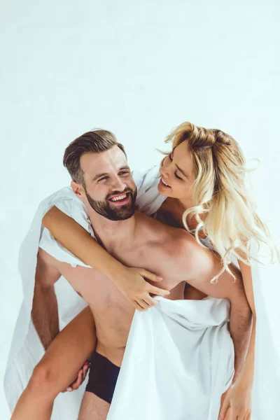 Cheerful young man piggybacking smiling girlfriend while wrapping in white sheet — Stock Photo