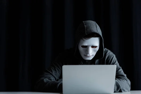 Анонимный интернет-тролль в маске, печатающий на клавиатуре ноутбука на черном — стоковое фото