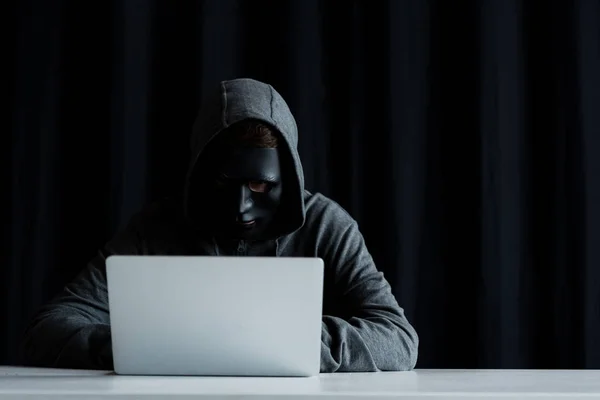 Anonymer Internet-Troll in Maske tippt auf Laptop-Tastatur auf schwarz — Stockfoto