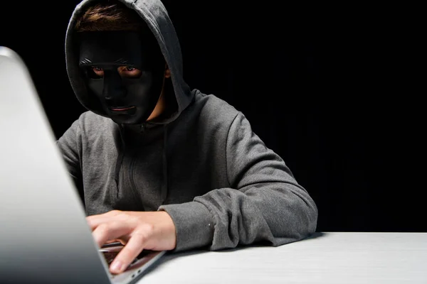 Anonymous internet troll in mask typing on laptop keyboard isolated on black — Stock Photo