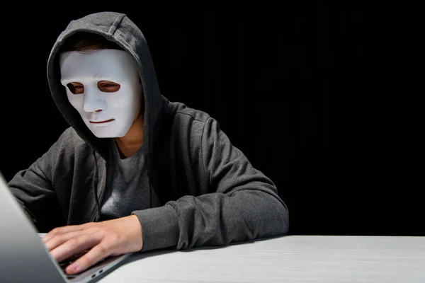Anonimo troll internet in maschera digitando su tastiera portatile isolato su nero — Foto stock