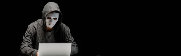 Панорамный снимок анонимного интернет-тролля в маске, печатающего на клавиатуре ноутбука, изолированного на черном — стоковое фото