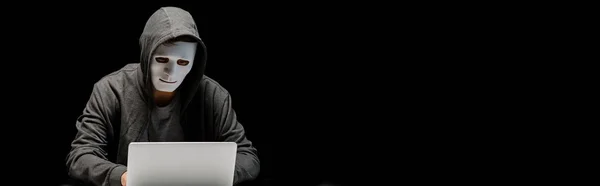 Panoramic shot of anonymous internet troll in mask typing on laptop keyboard isolated on black — Stock Photo