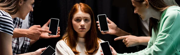 Panoramic shot of smiling teenagers pointing with fingers at girl during bullying and holding smartphones with blank screen on black — Stock Photo