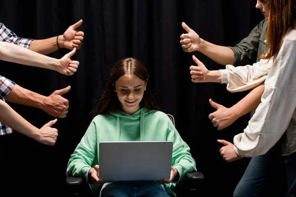 Vue partielle de personnes montrant pouces levés et souriant fille tenant ordinateur portable sur noir — Photo de stock