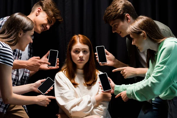 Smiling teenagers pointing with fingers at girl during bullying and holding smartphones with blank screen on black — Stock Photo