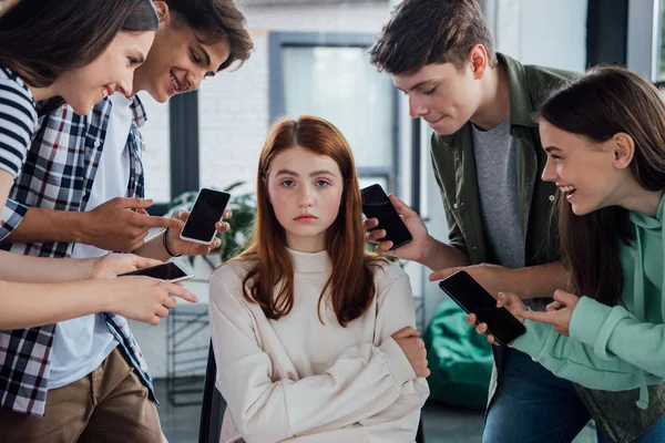 Adolescentes sonrientes señalando con los dedos a la chica durante el acoso escolar y sosteniendo teléfonos inteligentes con pantalla en blanco - foto de stock