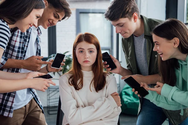 Lächelnde Teenager zeigen beim Mobbing mit den Fingern auf Mädchen und halten Smartphones mit leerem Bildschirm — Stockfoto