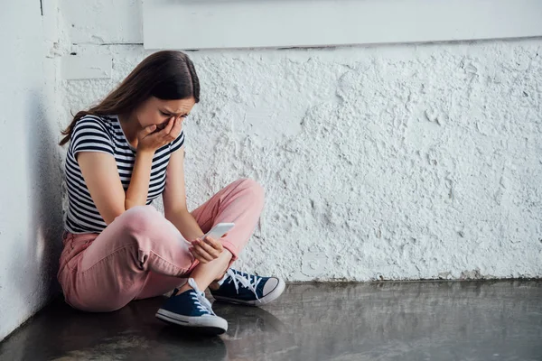 Trauriges weinendes Mädchen in pinkfarbener Hose, das neben Wand sitzt und Smartphone in der Hand hält — Stockfoto