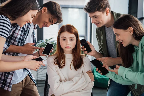 Улыбающиеся подростки, указывающие пальцами на девушку во время издевательств и держащие смартфоны с пустым экраном — стоковое фото
