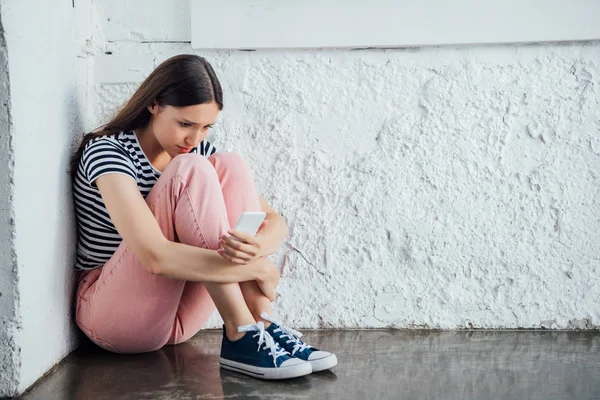 Menina triste em calças rosa sentado perto da parede e segurando smartphone — Fotografia de Stock