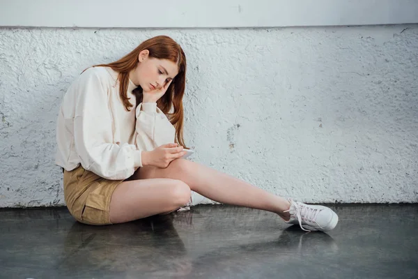 Sad girl sitting near textured wall and holding smartphone — Stock Photo