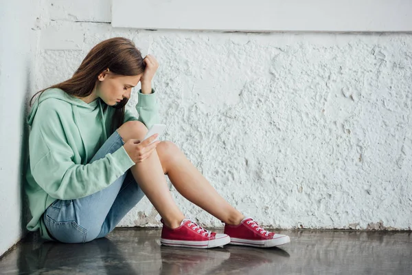 Sad girl sitting near textured wall and holding smartphone — Stock Photo