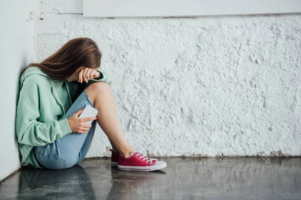 Chica llorando triste sentada cerca de la pared texturizada y sosteniendo teléfono inteligente - foto de stock