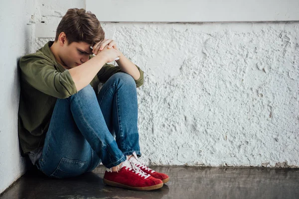 Triste adolescente en jeans celebración de teléfono inteligente y sentado en el suelo - foto de stock