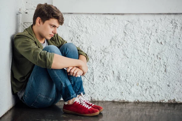 Грустный подросток в джинсах держит смартфон и сидит на полу — стоковое фото