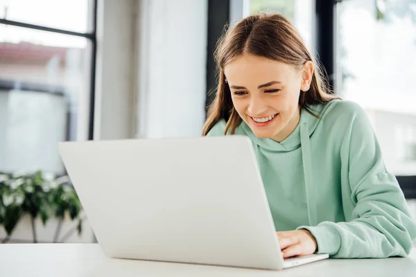 Sonriente chica en casual sudadera con capucha sentado en la mesa y el uso de ordenador portátil - foto de stock