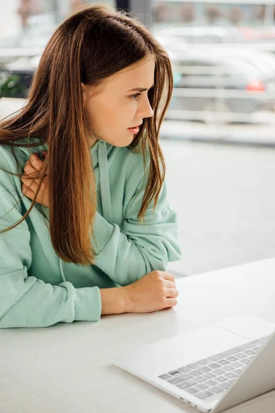 Сумна дівчина в повсякденному светрі сидить за столом і використовує ноутбук — стокове фото